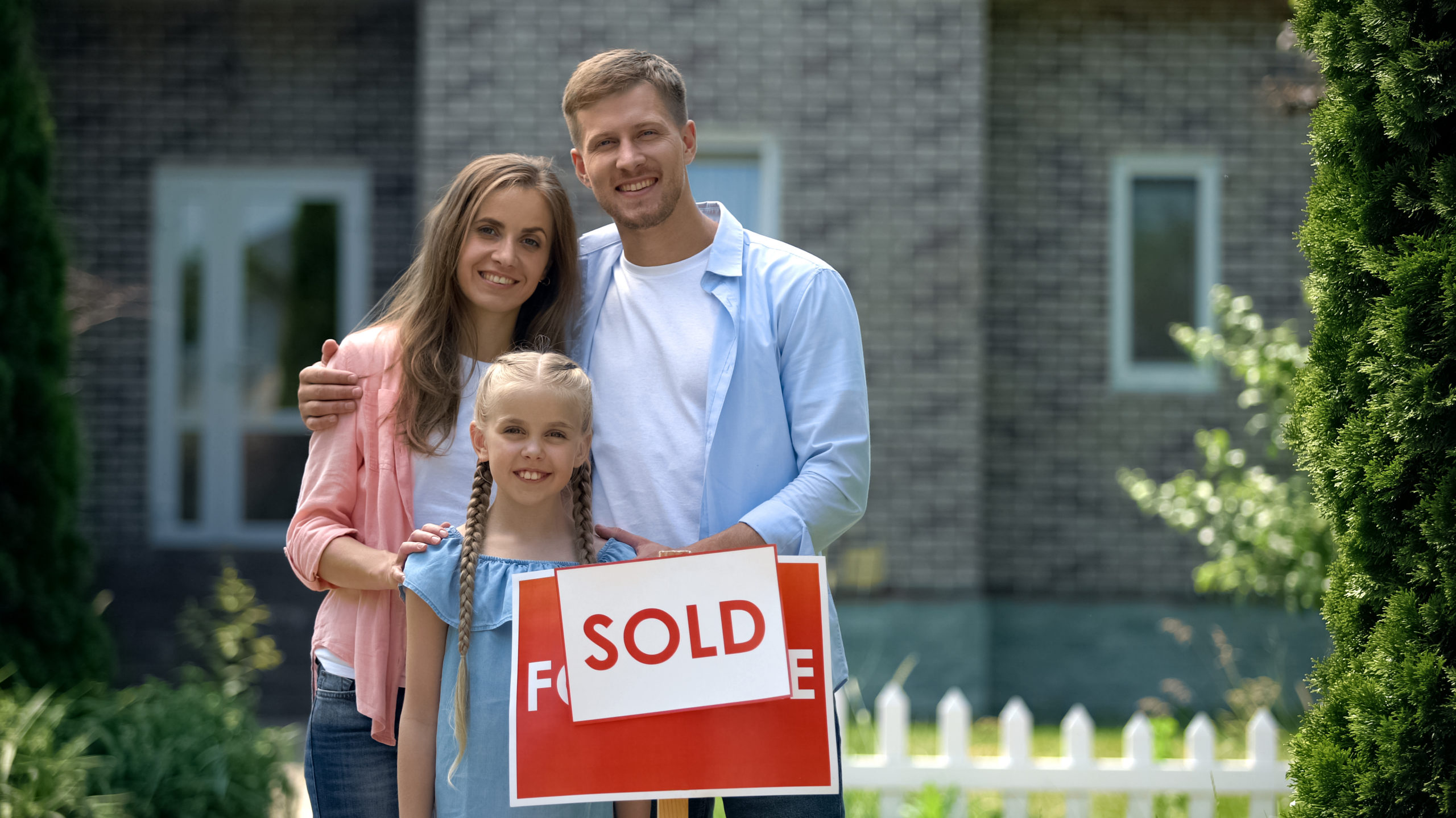 Happy family holding sold sign, standing against new bought house, thumbs up